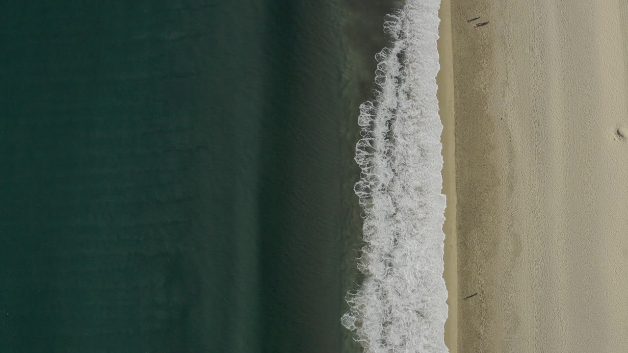 Overhead frame of a Baja California Sur beach where dark blue-green water meets pale sand through a vertical band of white foam, with no visible people.