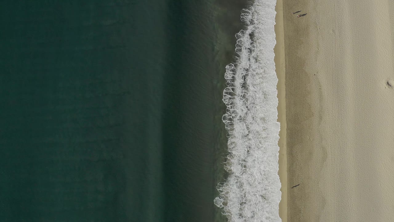 Aerial view over Baja California Sur shoreline with the foam line shifted along the beach, deep water on the left, pale sand on the right, and no visible people.