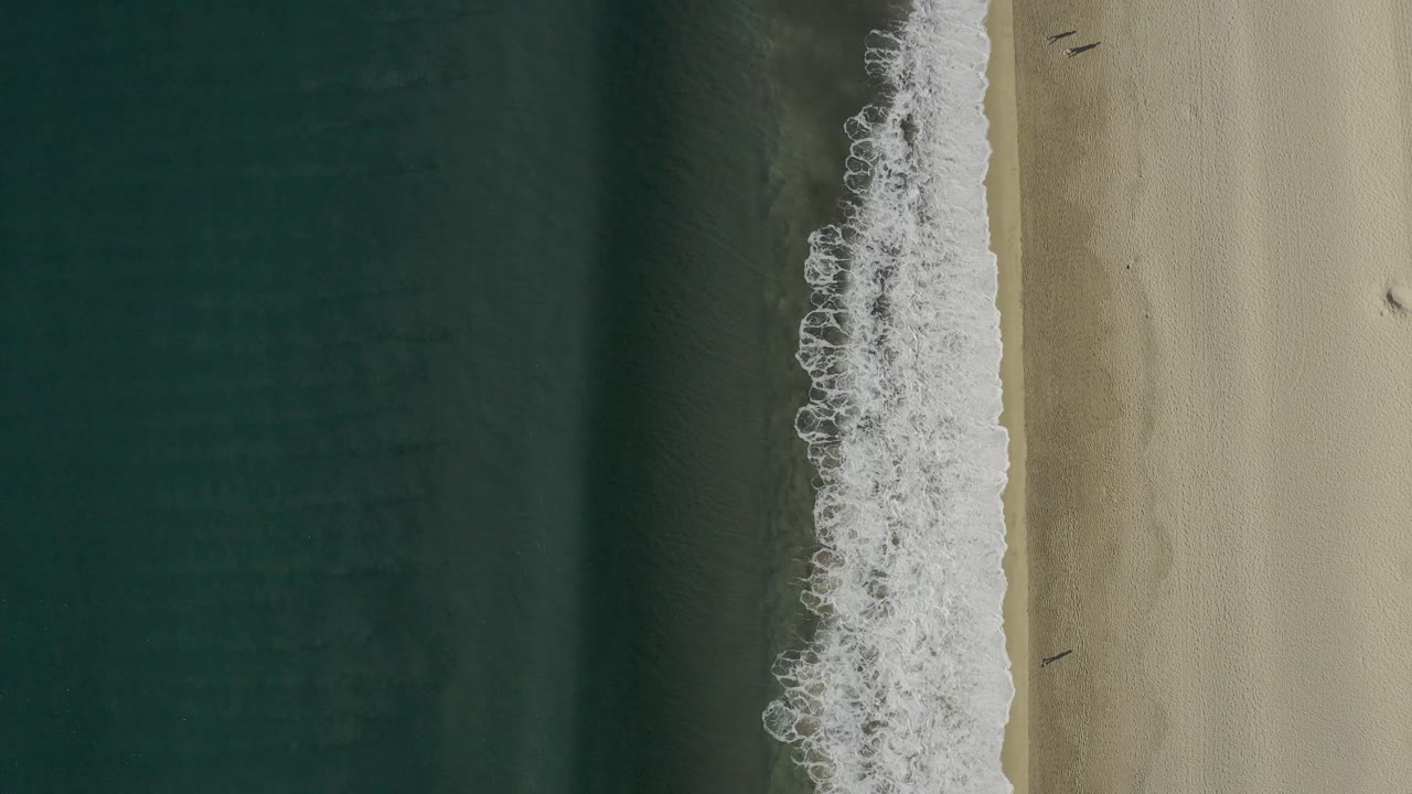 Top-down Baja California Sur beach frame with a narrow white surf band, darker shallow water to the left, smooth light sand to the right, and no visible people.