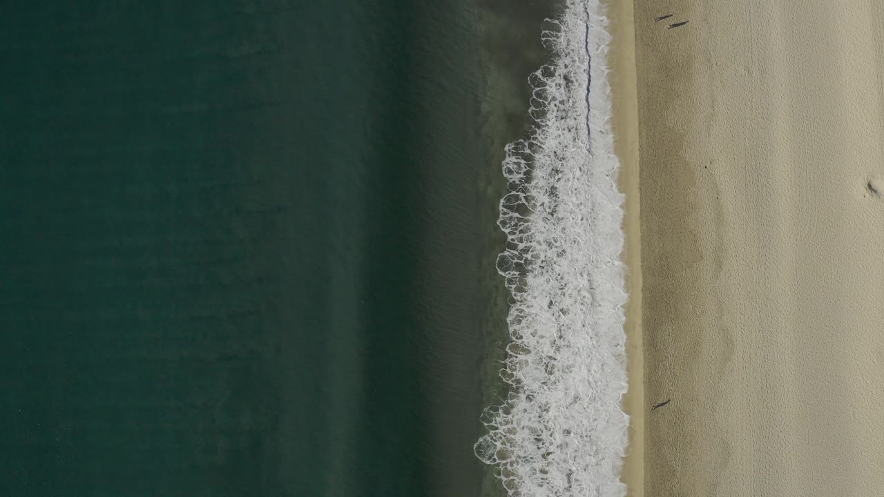 Aerial frame from Baja California Sur showing white foam spreading more broadly along the vertical shoreline, with blue-green water and pale beach visible and no visible people.