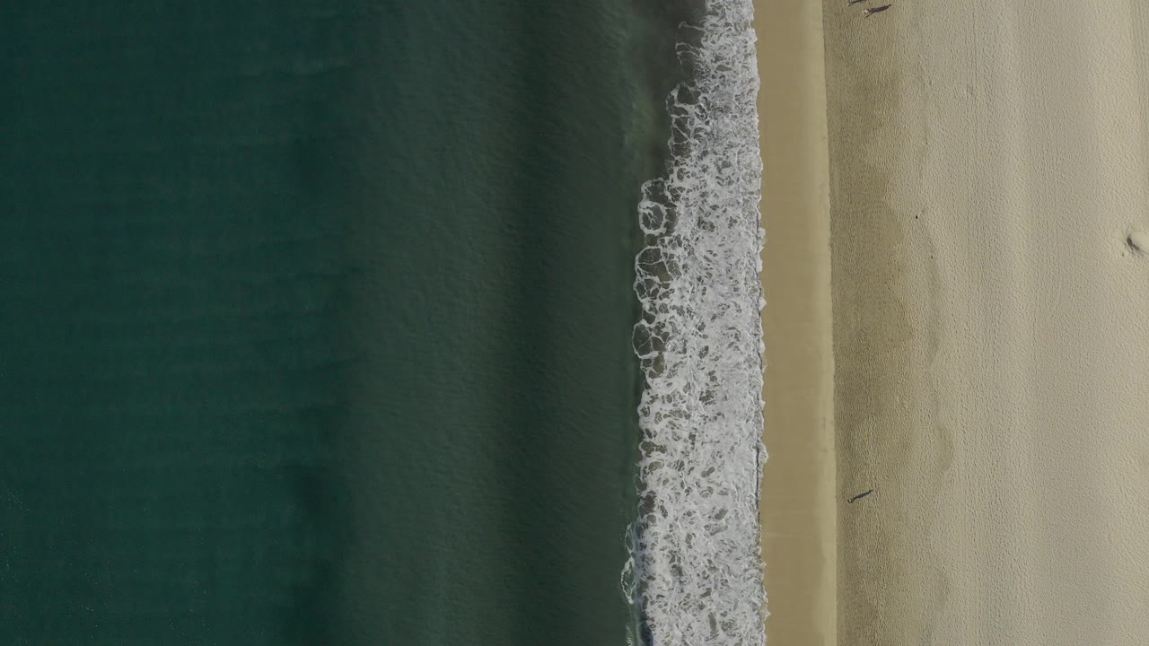 Overhead shoreline with blue-green water, white surf, and pale sand.
