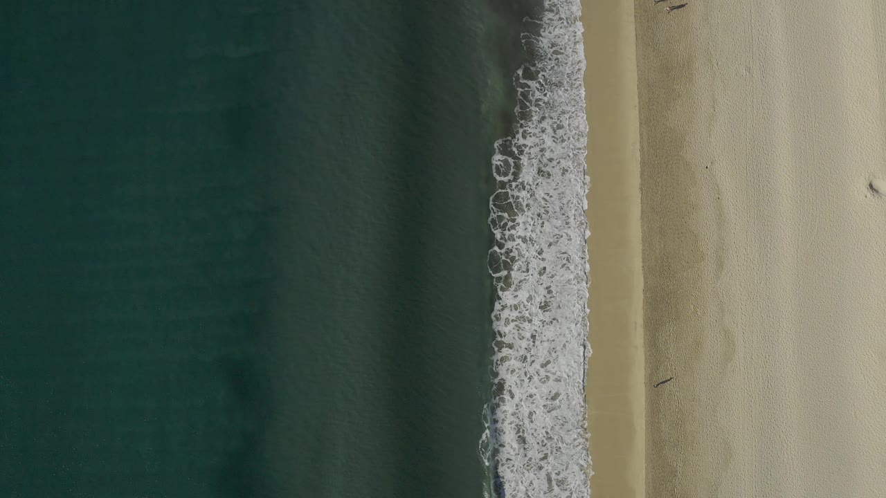 Aerial frame of Baja California Sur beach where a thicker band of white wave foam presses against wet sand, with open water to the left and no visible people.