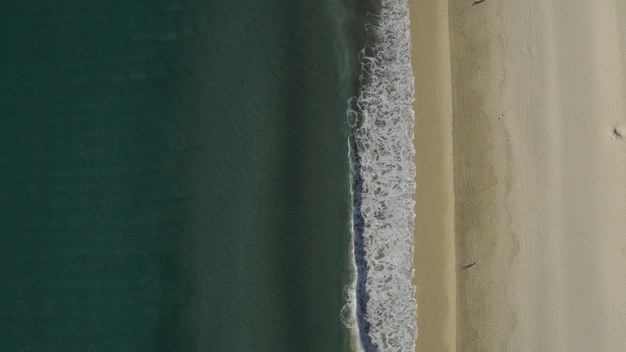 Top-down view of the Baja California Sur coast with dark blue-green water, a textured white surf edge, and empty pale beach to the right, no visible people.