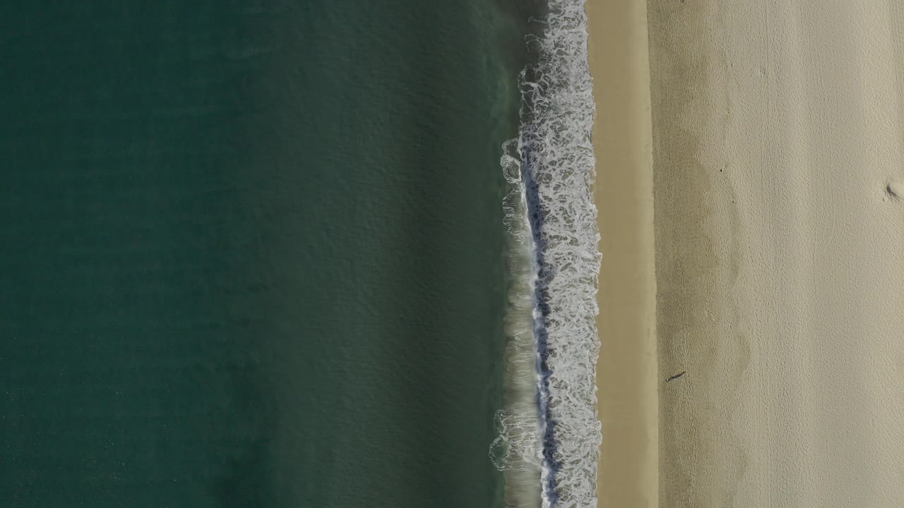 Direct overhead frame in Baja California Sur showing foamy surf sliding along a vertical shoreline between green water and pale sand, no visible people.