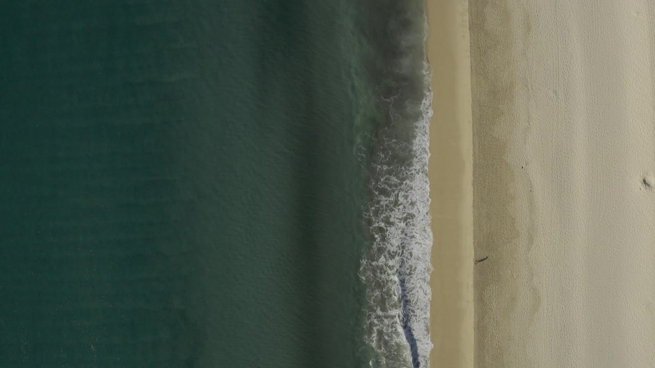 Top-down aerial frame of the Baja California Sur shoreline with dark teal water at left, a thin white surf edge, pale sand at right, and no visible people.
