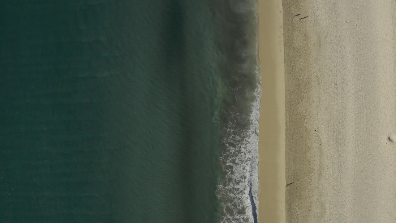 Straight-down frame over Baja California Sur showing vertical bands of dark water, narrow surf, and broad pale sand with no visible people.