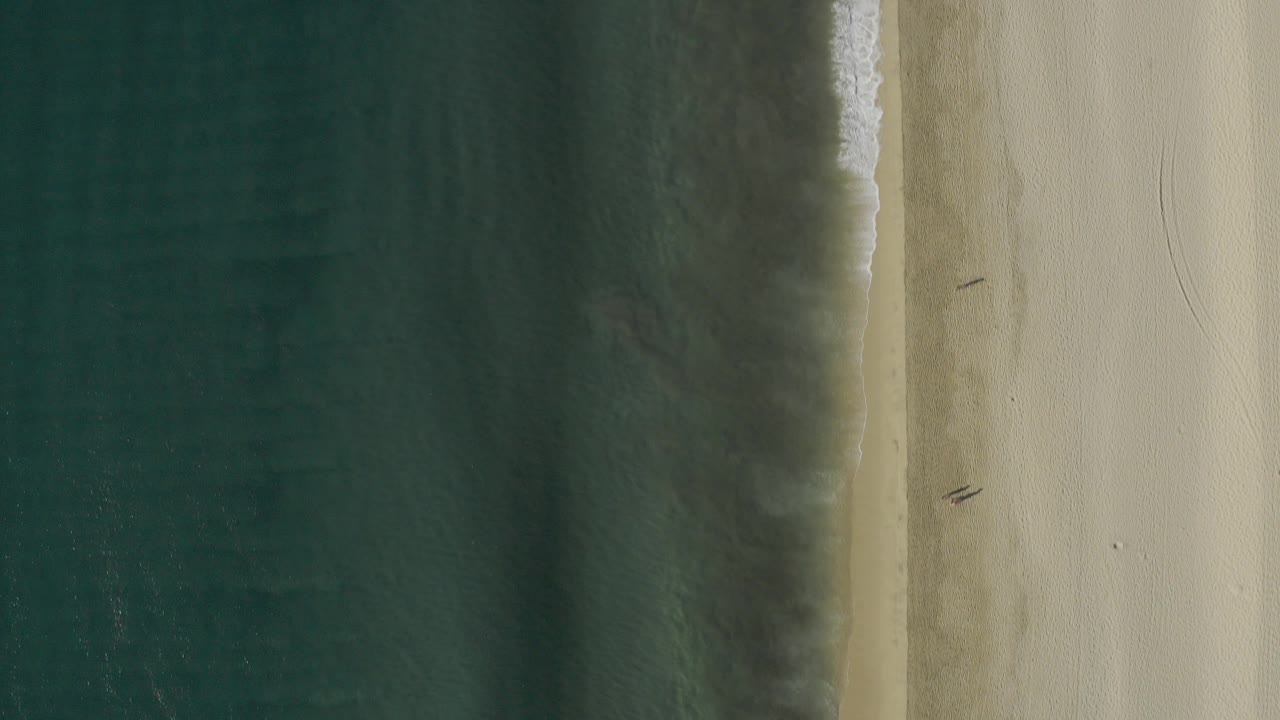 Top-down aerial frame over Baja California Sur, Mexico, with dark green-blue water on the left, a thin breaking wave line, pale sand on the right, and no visible people.