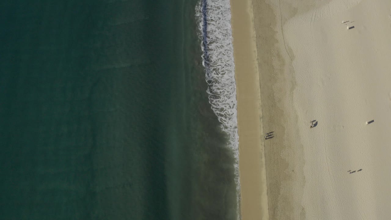Top-down view of Baja California Sur ocean meeting tan beach at the start of the clip, with dark water on the left, dry sand on the right, and no visible people.