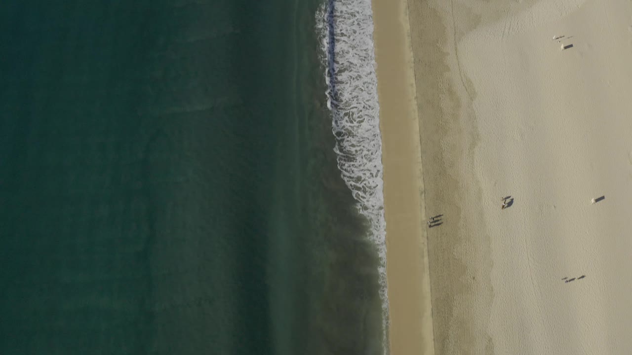 Aerial frame over Baja California Sur coastline with the surf line running vertically through the shot, faint beach tracks in the sand, and no visible people.
