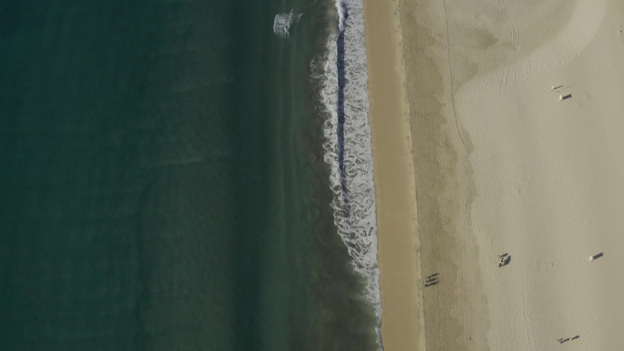 Top-down Baja California Sur beach frame with small white waves breaking near the sand, darker water bands offshore, and no visible people.