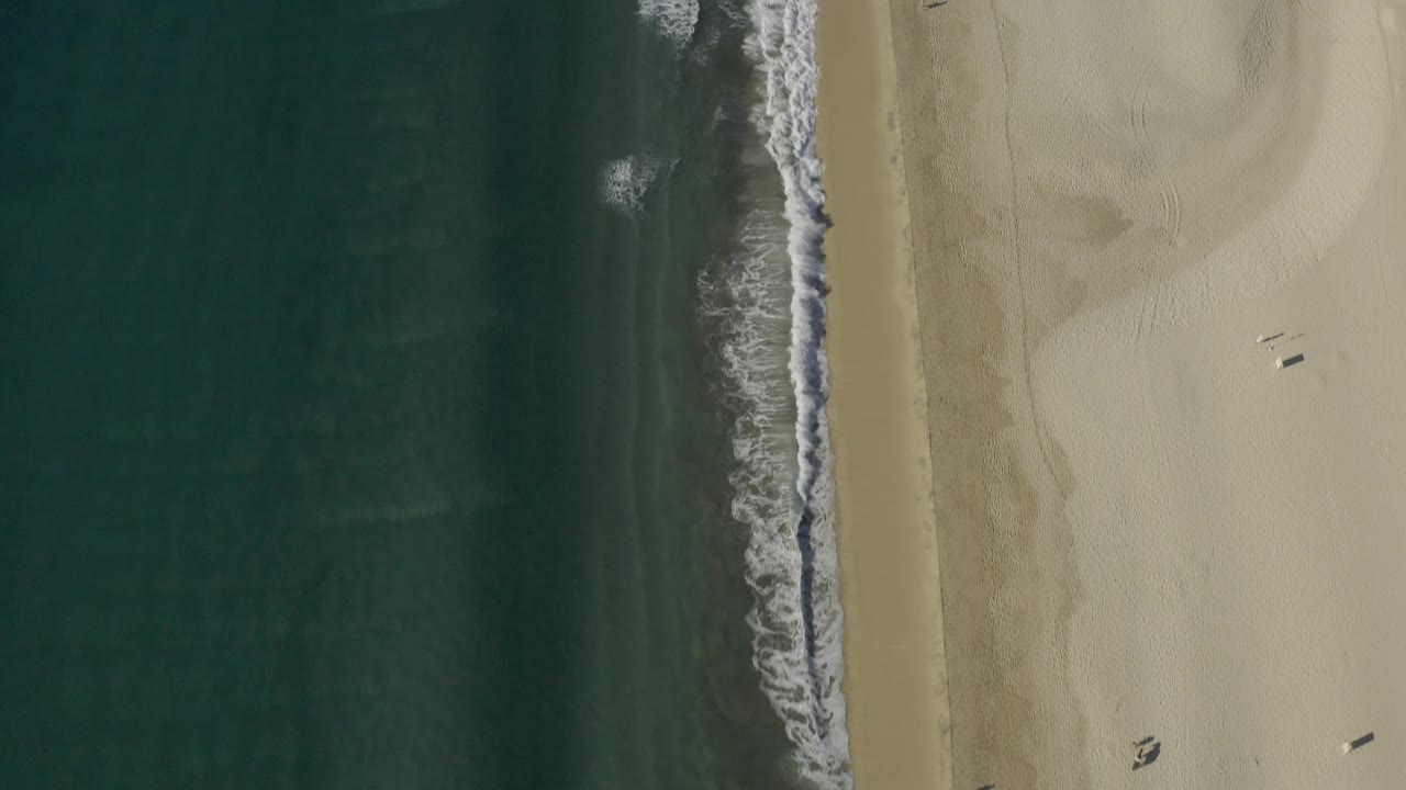 Overhead coastline view in Baja California Sur with a narrow foamy wave line, broad empty tan sand to the right, and no visible people.