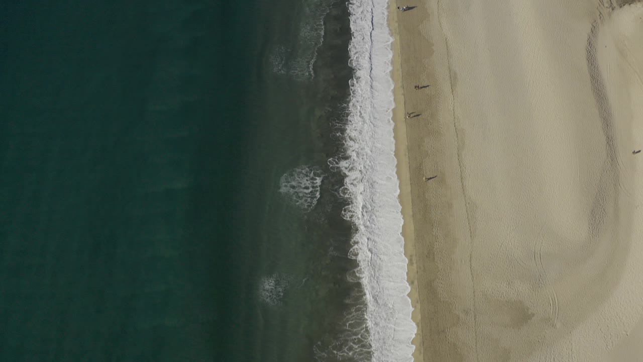 Overhead view in Baja California Sur Mexico with dark green water, a white surf edge, pale sand, and no visible people at the start of the clip.