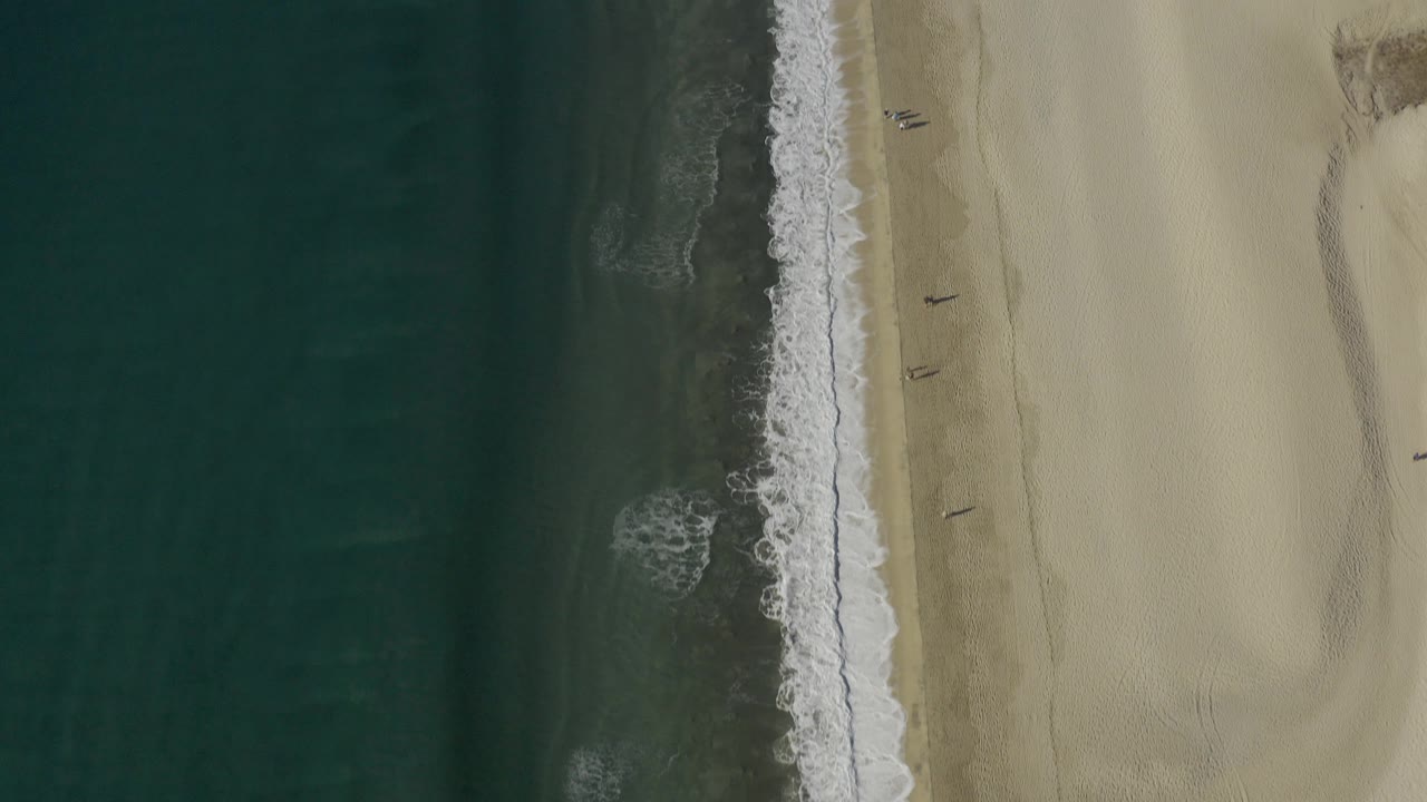 Top-down Baja California Sur Mexico shoreline frame with darker water areas, foamy surf down the center, pale beach to the right, and no visible people.
