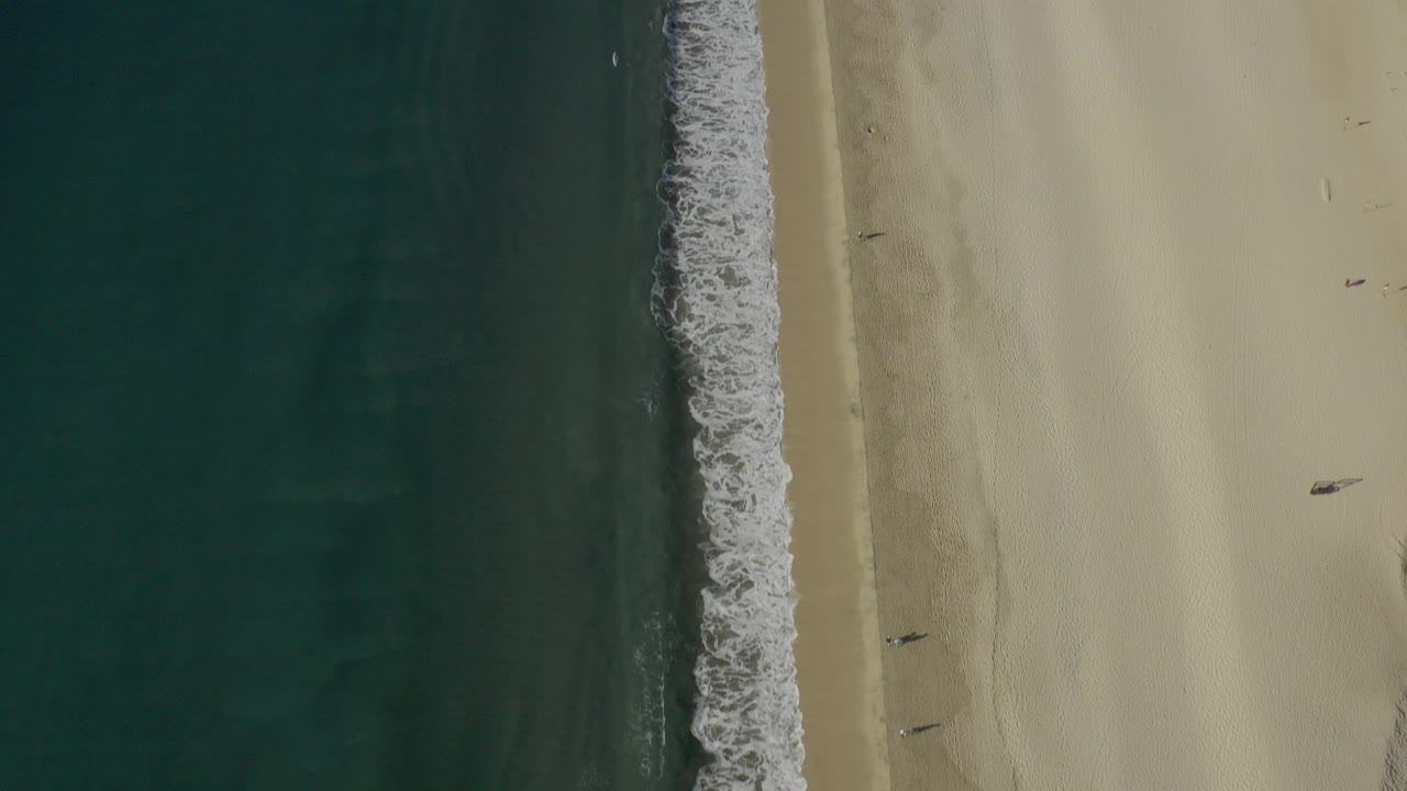 Overhead view of a Baja California Sur beach with dark teal water on the left, a foamy surf edge, pale sand, and several tiny people visible along the shore.