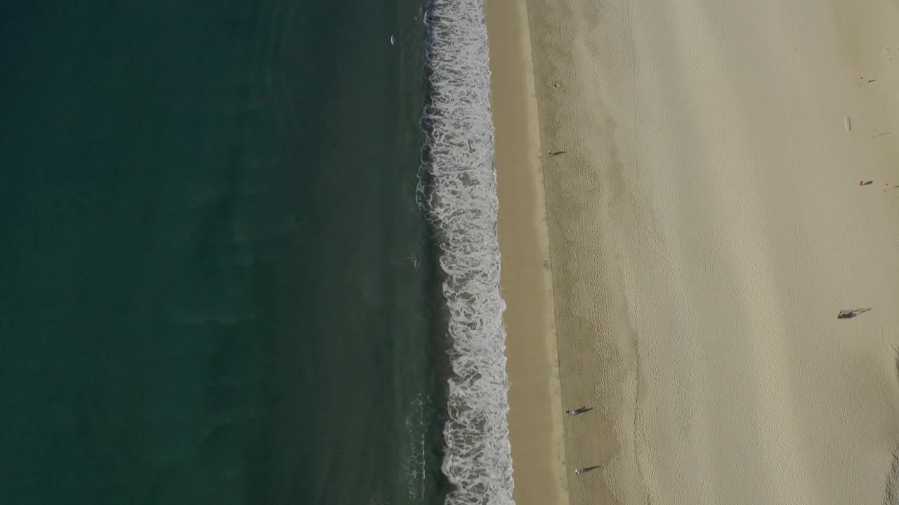 Aerial beach line with surf beside open sand and a few tiny figures.