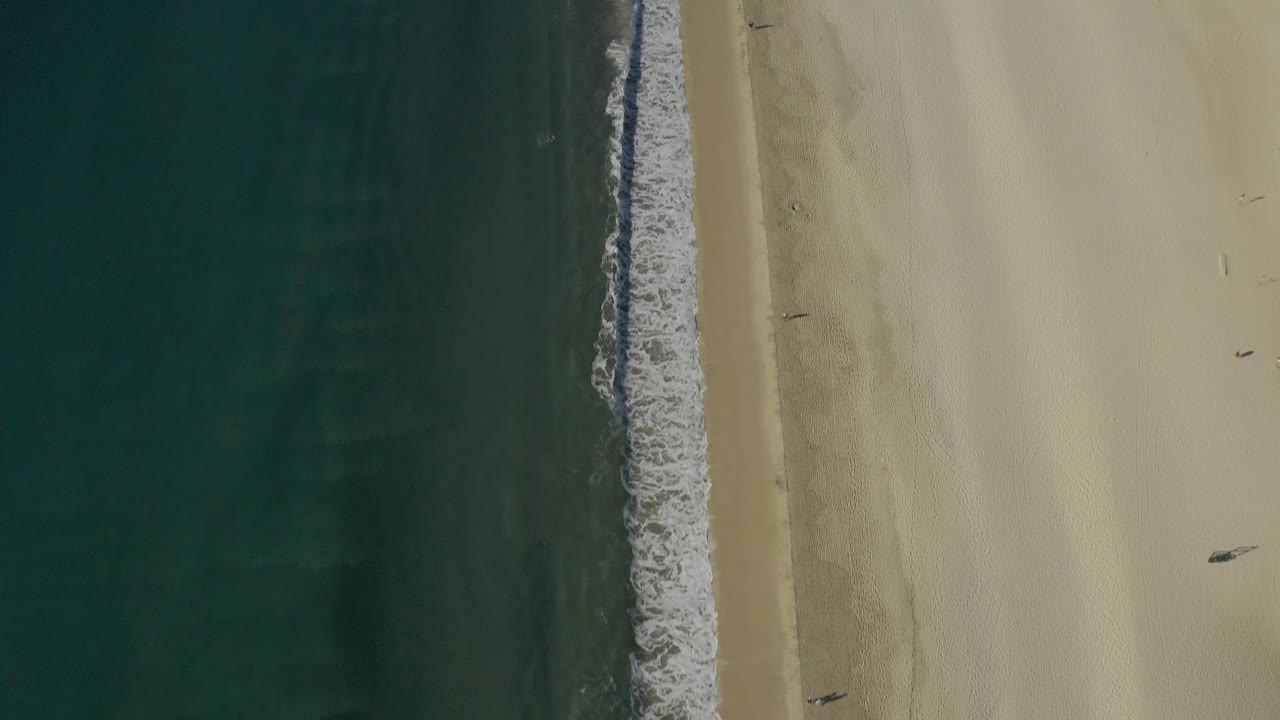 Top-down view of the Baja California Sur coast with deep green-blue water, a narrow white breaker, pale beach, and scattered tiny people visible on the sand.