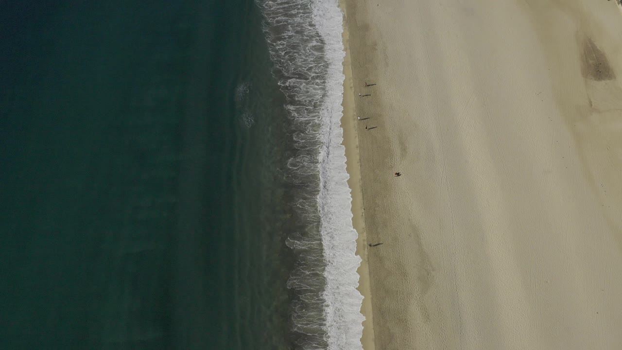 Overhead coastline with dark ocean, white surf, and pale sand.