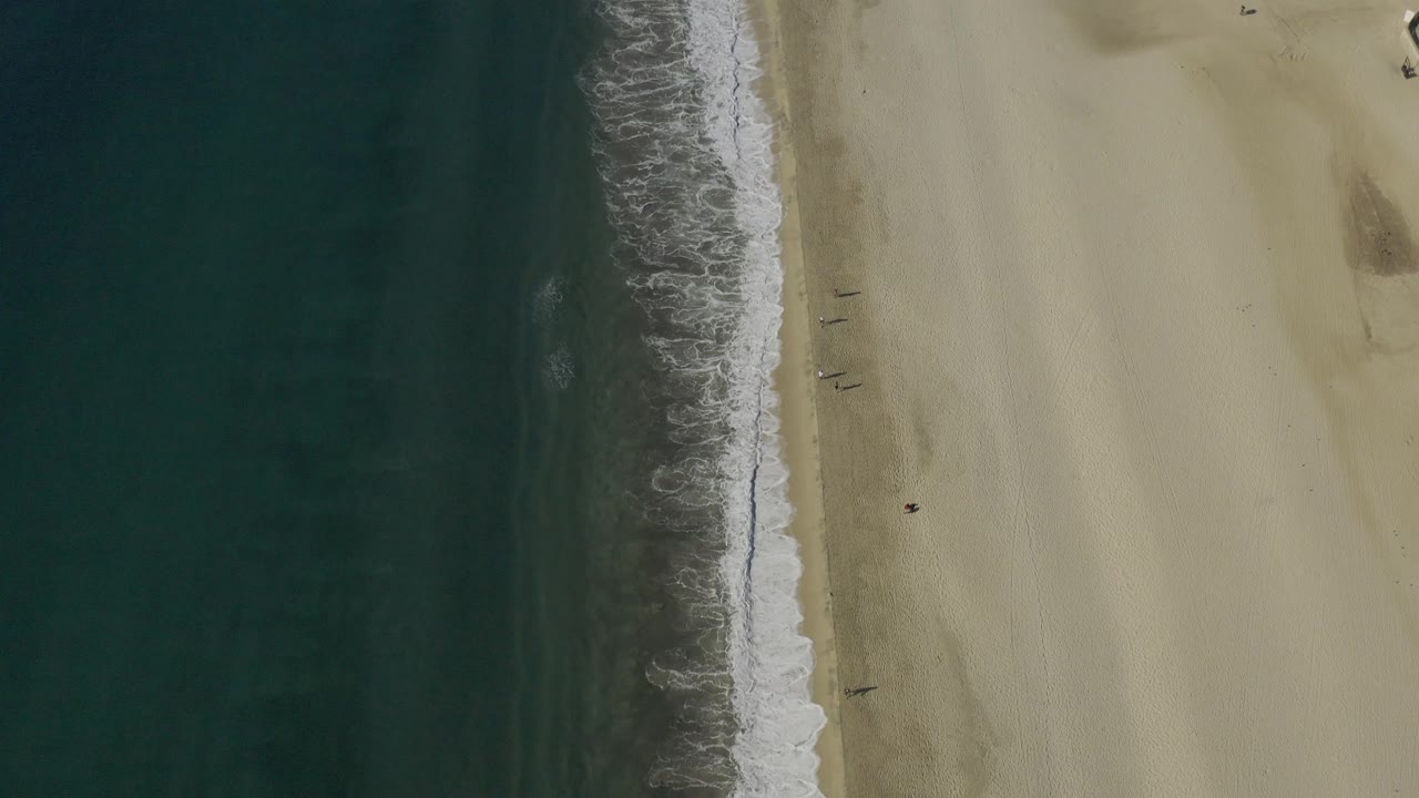 Aerial frame from Baja California Sur showing foamy surf running down the center, darker ocean water to the left, sunlit sand to the right, and no visible people.