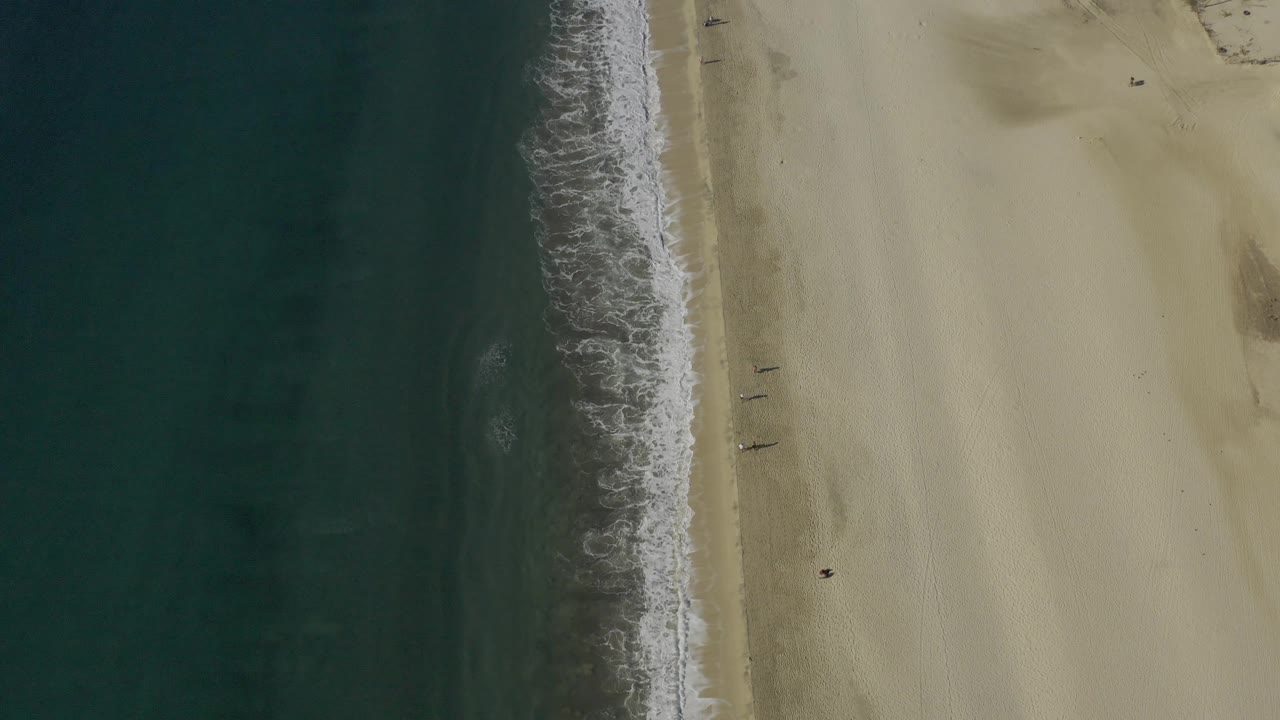 Overhead Baja California Sur beach view with deep green-blue water on the left, a ragged white wave band near center, pale sand on the right, and no visible people.