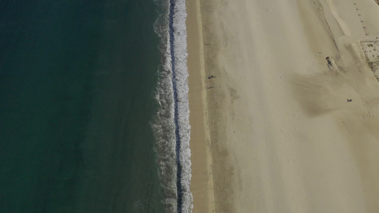Top-down aerial frame from Baja California Sur, Mexico, with dark blue-green water on the left, a narrow surf line, pale sand on the right, and no visible people.