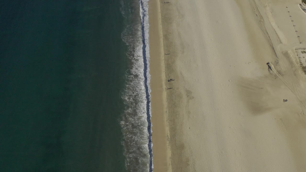 Top-down Baja California Sur shoreline frame with dark ocean occupying the left half, a narrow breaking wave near center, pale sand to the right, and no visible people.
