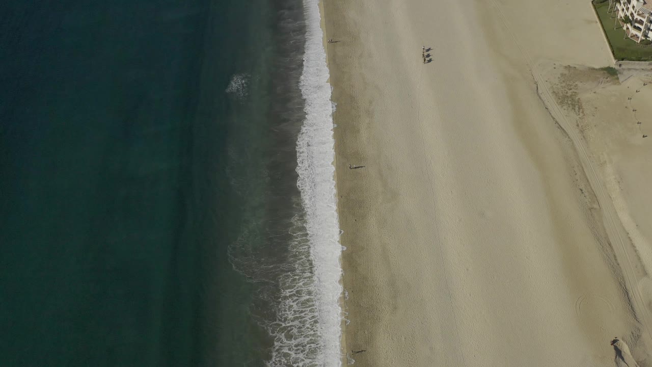 Aerial view in Baja California Sur showing the surf line slightly lower in frame, with dark water on the left, open sand on the right, and no visible people.