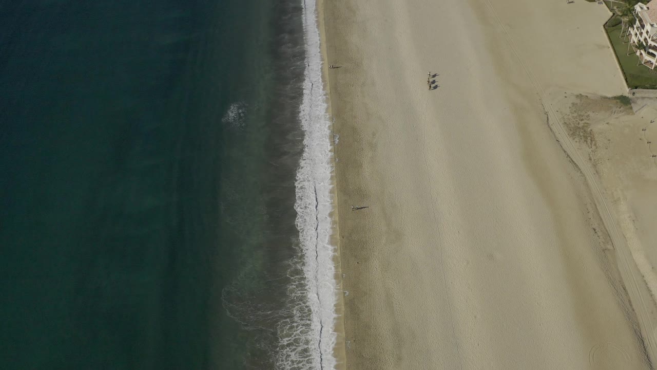 Drone-like overhead frame of the Baja California Sur coast with dark water, a thin white surf strip, pale sand, and small structures at the upper right, with no visible people.