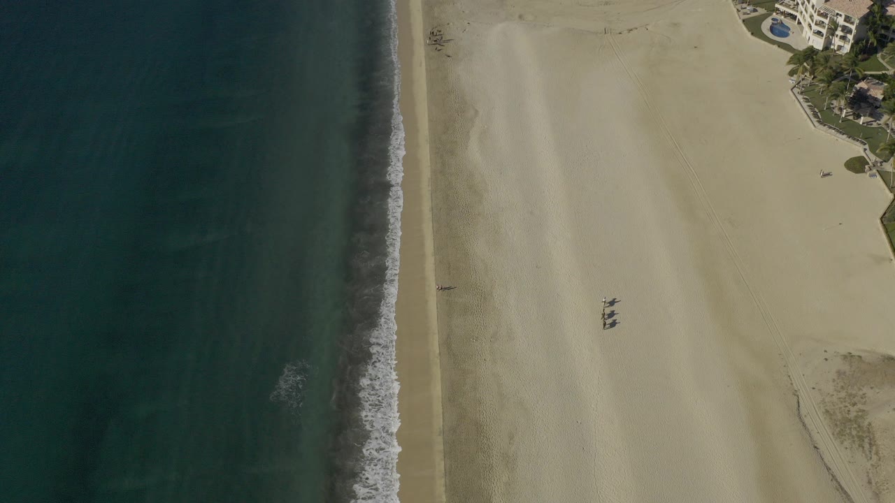 Overhead view of Baja California Sur coastline with dark ocean at left, pale beach at center, beachfront apartment buildings at upper right, and no visible people.