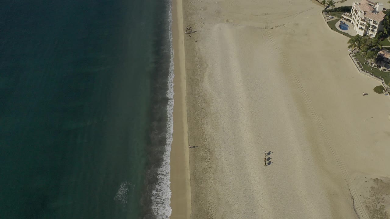 Drone-style overhead frame of Baja California Sur coastline with darker water bands offshore, a narrow foamy wave edge, open sand, and apartment buildings at top right with no visible people.