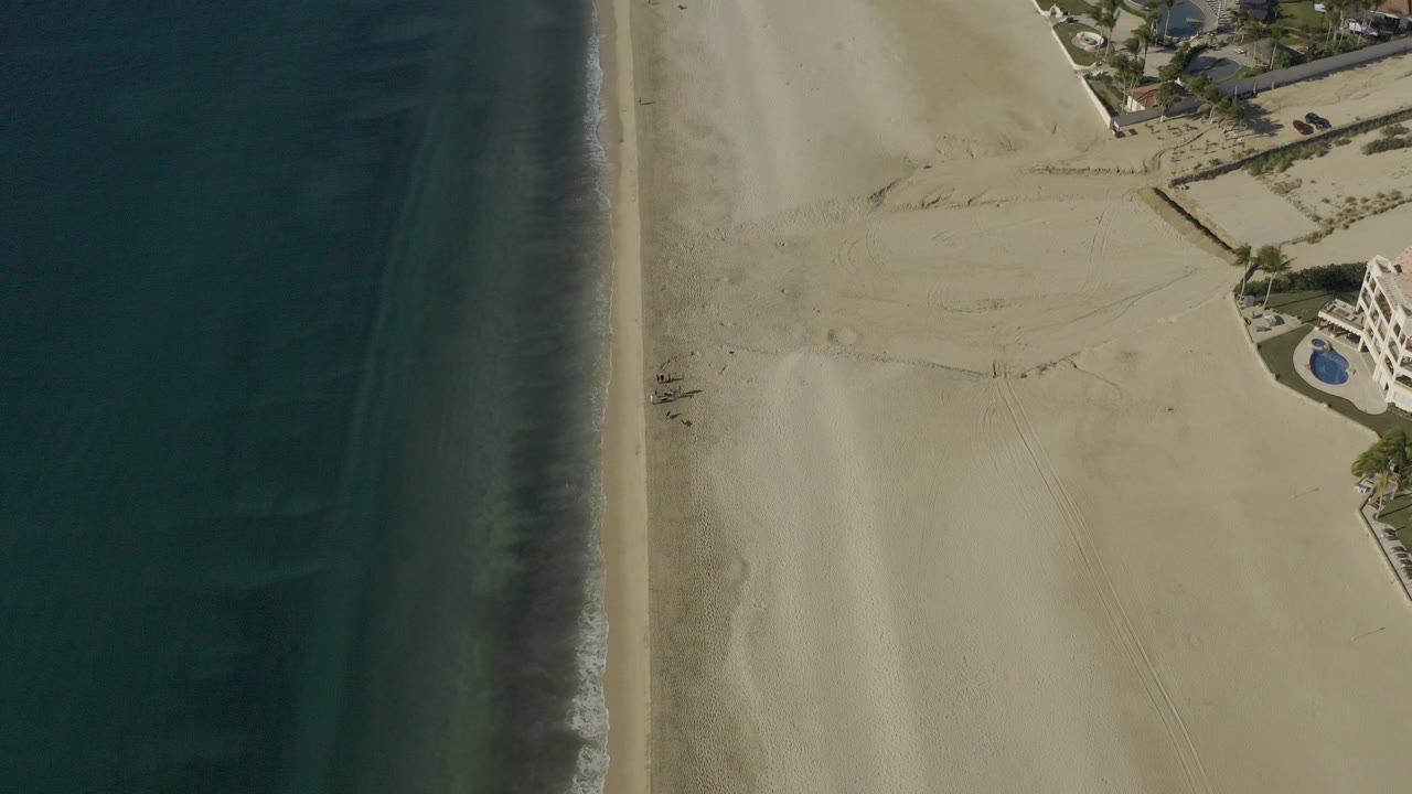 High overhead view in Baja California Sur, Mexico showing dark ocean at left, a thin surf edge, broad pale beach, and beachfront buildings with pools near the upper right, with no visible people.