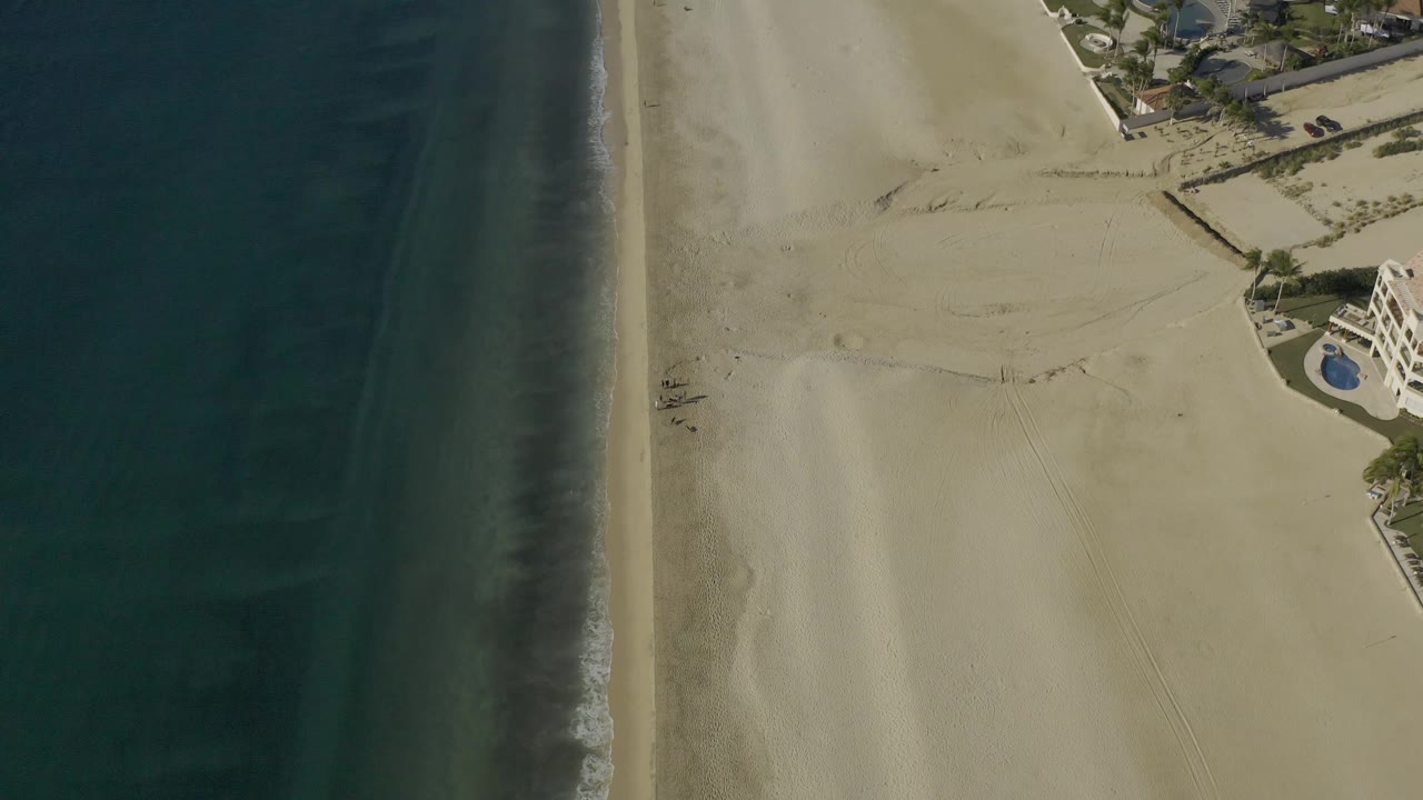 Aerial frame from Baja California Sur, Mexico with dark water filling the left side, a straight narrow shoreline, open sand in the middle, and poolside beachfront structures cropped at the right edge, with no visible people.