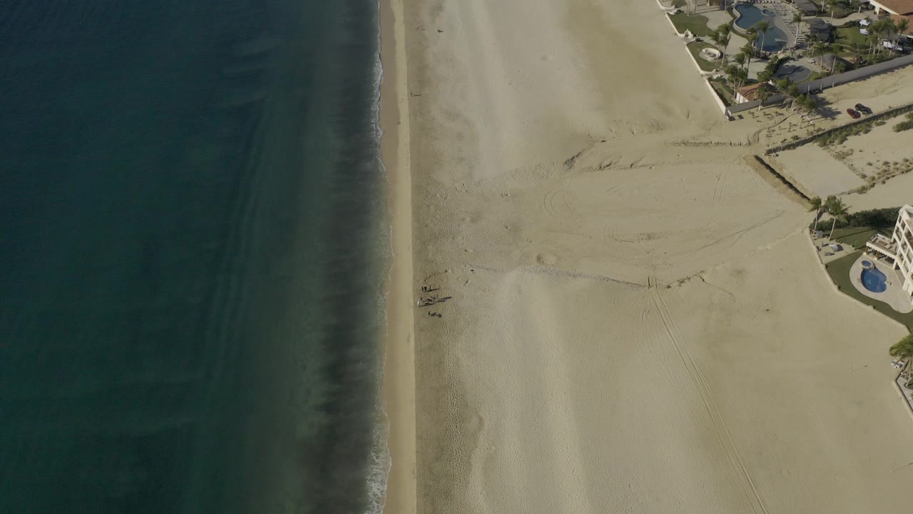 High drone-style view in Baja California Sur, Mexico showing the ocean along the left third, smooth sand across the center, darker beach marks near the waterline, and homes or resort buildings with blue pools at the upper right, with no visible people.