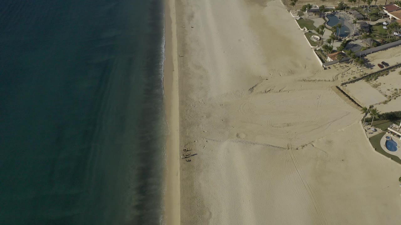 Coastline from above with dark water, pale surf edge, sandy beach, and nearby pool areas