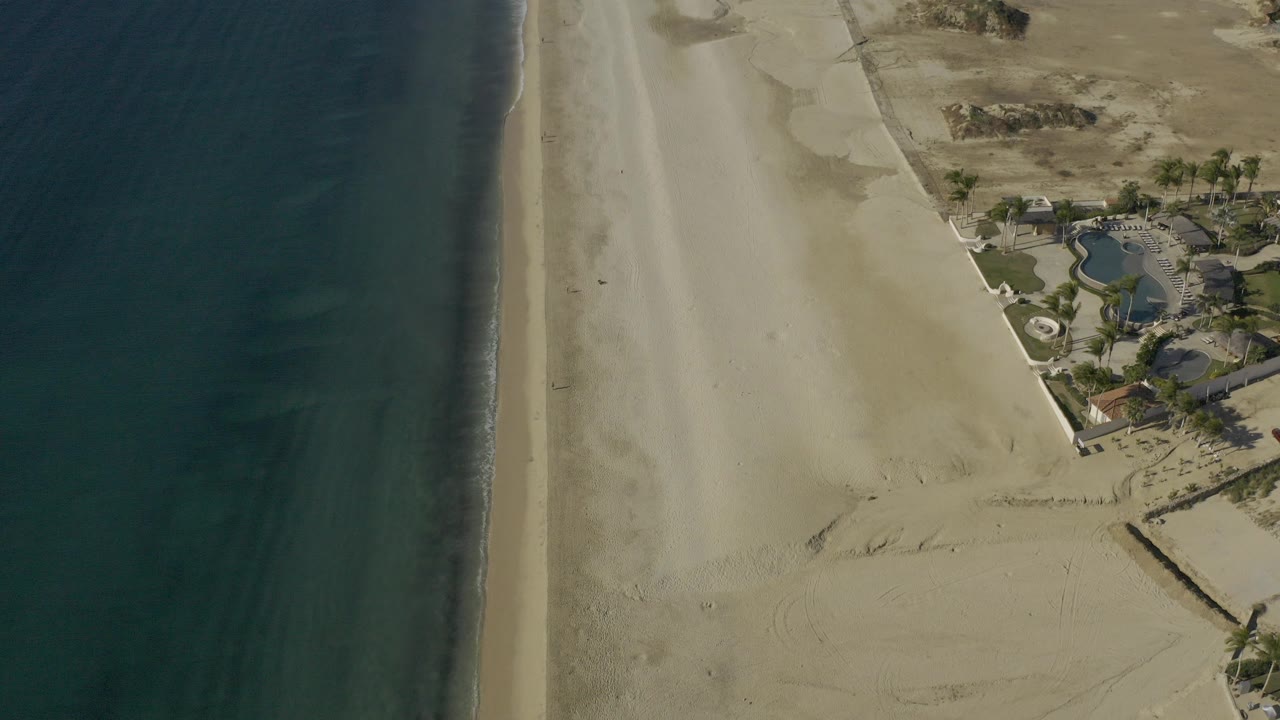 High aerial view in Baja California Sur, Mexico with dark ocean at left, wide pale beach through the center, dunes and beachfront buildings at right, and no visible people.