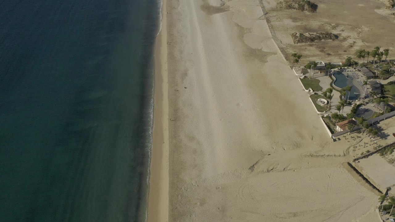 Aerial shoreline frame in Baja California Sur, Mexico showing blue-green water, a narrow surf edge, wide sand, dry dunes, curved landscaped property, and no visible people.