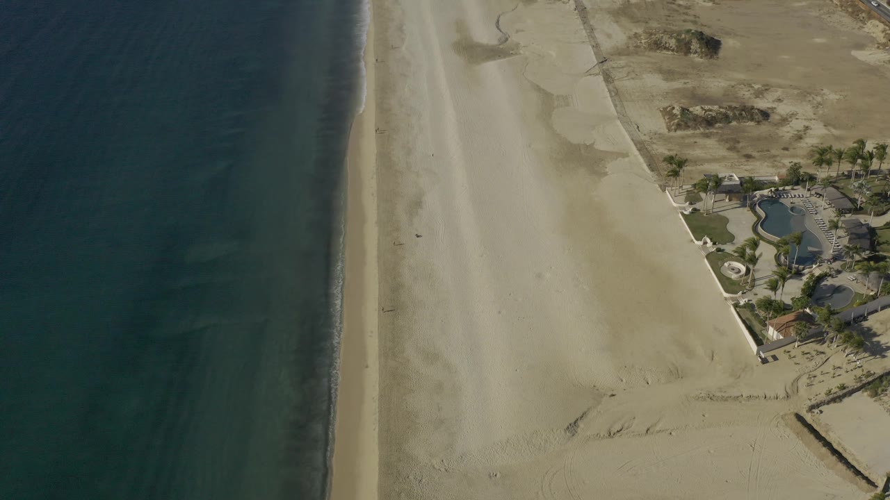 Baja California Sur aerial frame with ocean and foamy shoreline on the left, a broad sandy beach, dry open land, and beachfront buildings near the upper right, with no visible people.