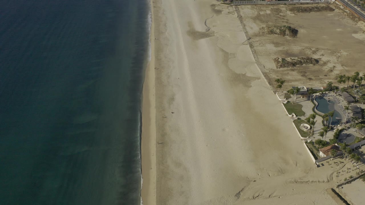 Aerial coastline with dark water, pale sand, dune patches, and landscaped buildings.