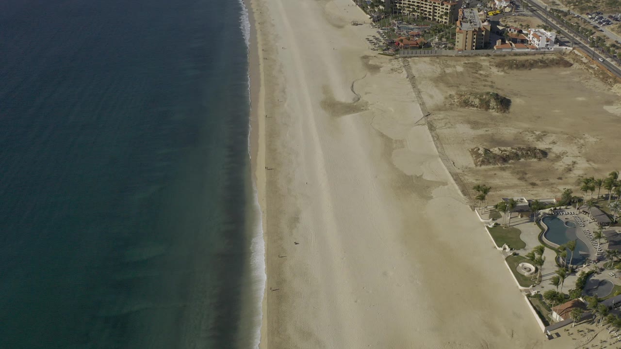Aerial frame over Baja California Sur coastline with dark blue water on the left, pale sand beach in the middle, dry dunes and buildings to the right, and no visible people.