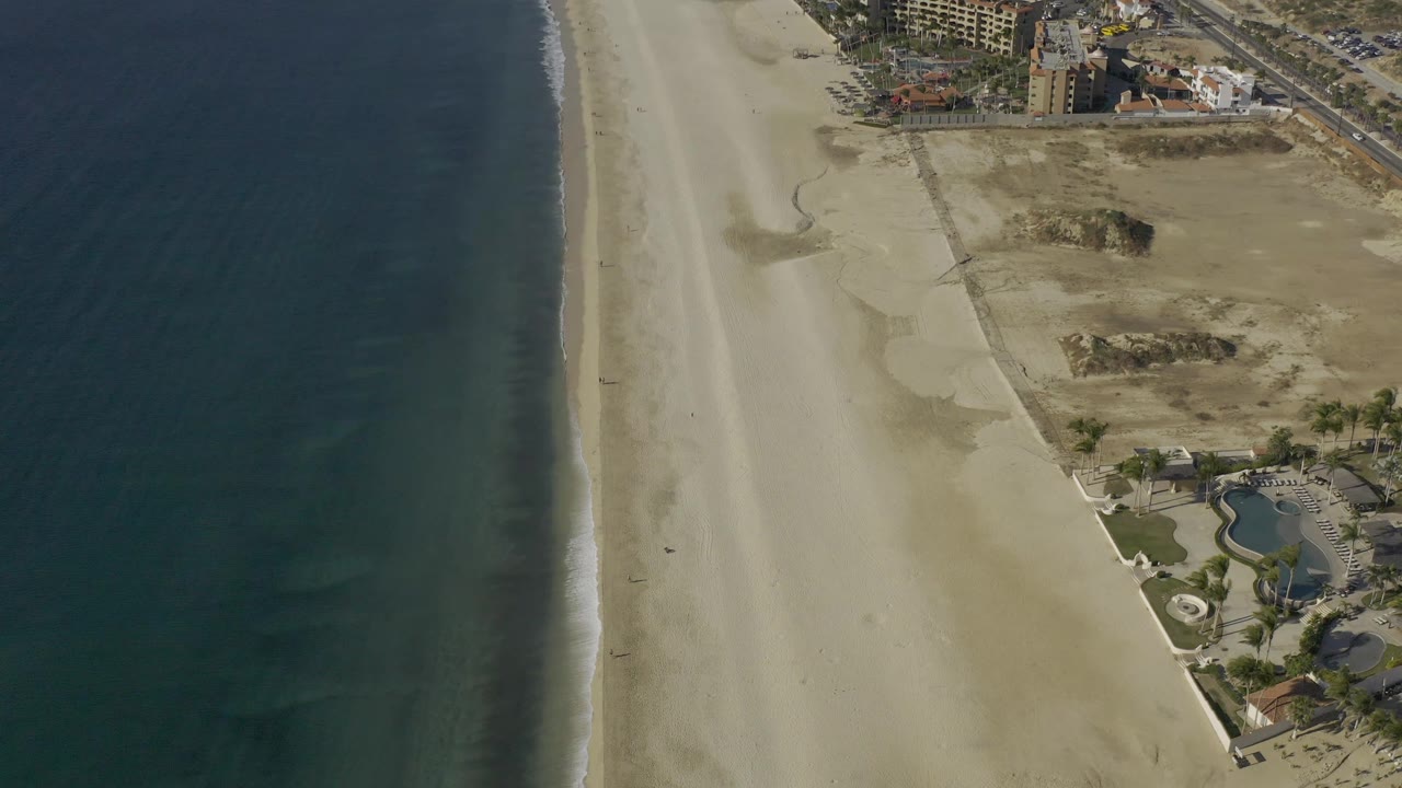 Overhead view of Baja California Sur beach with the surf line slightly farther left, open sand through the center, dry inland lots, resort-like buildings and pools on the right, and no visible people.