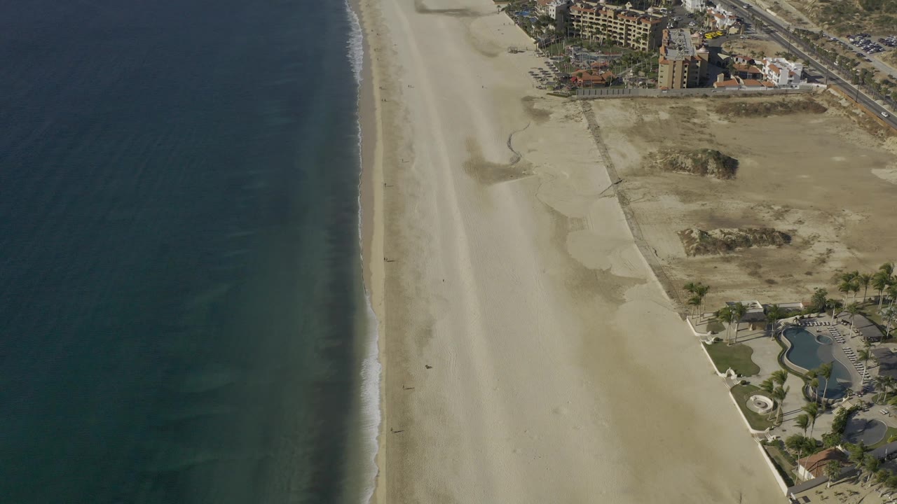 High aerial frame of the Baja California Sur shoreline with deep blue water, a narrow white wave edge, wide empty sand, dune patterns, and clustered buildings near the upper right, with no visible people.