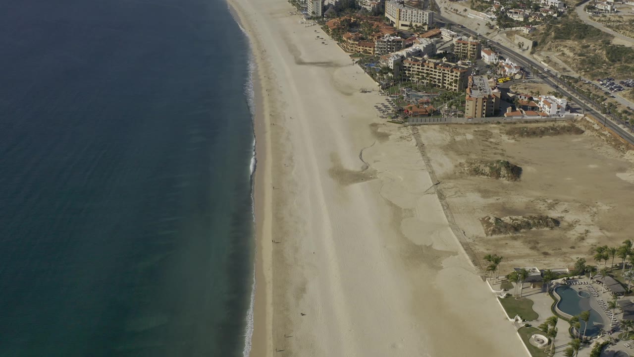 Aerial view in Baja California Sur, Mexico with the surf line running beside a broad sand beach, beachfront buildings near the upper right, and no visible people.