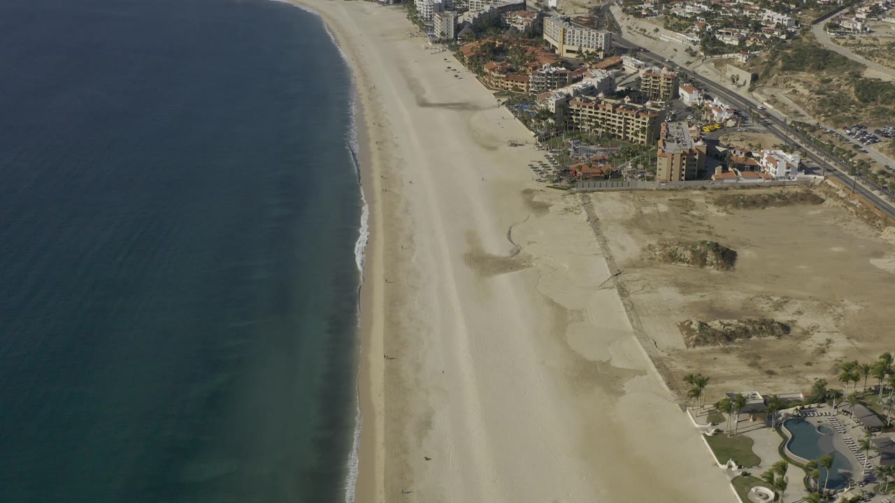 High aerial frame over Baja California Sur, Mexico showing a long empty beach beside dark water, with roads, parked cars, buildings, and dry lots inland, with no visible people.