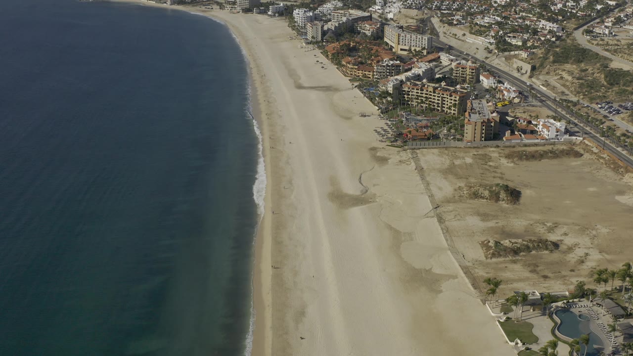 Aerial frame in Baja California Sur, Mexico with ocean filling the left side, a pale beach running vertically through frame, and resort-style buildings and open lots on the right, with no visible people.