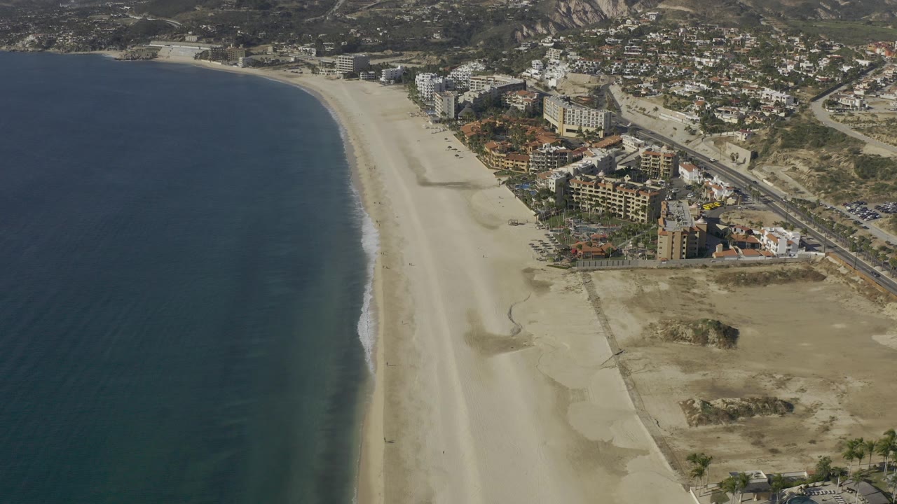 Aerial view over Baja California Sur with dark blue water, a broad pale sand beach, shoreline buildings, roads, dry hills, and no visible people.