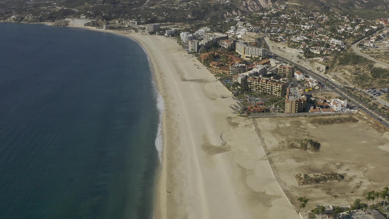 High aerial frame in Baja California Sur showing the ocean along the left edge, empty sand through the center, coastal buildings and roads to the right, and no visible people.