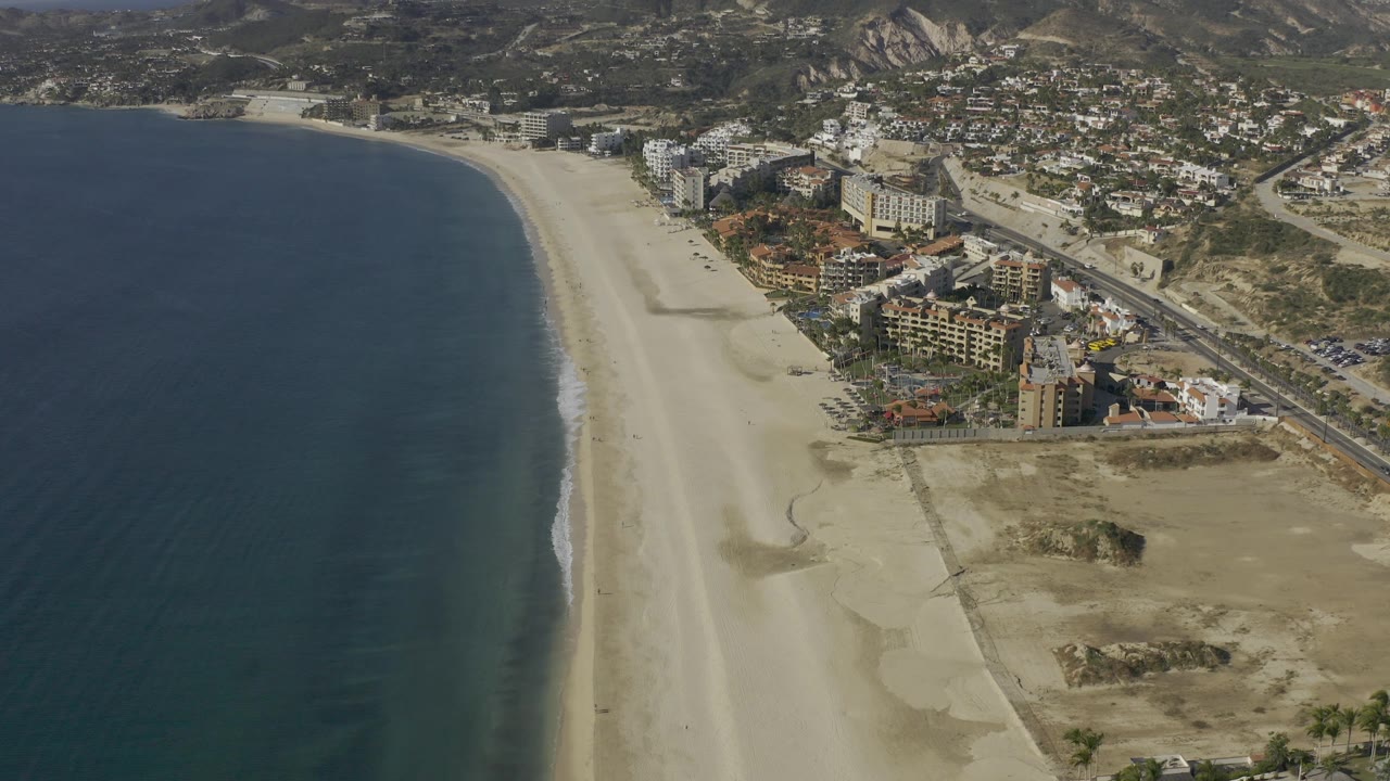 Drone-like aerial over Baja California Sur with a long empty beach, calm dark water, a row of coastal buildings, a road corridor, rugged hills, and no visible people.