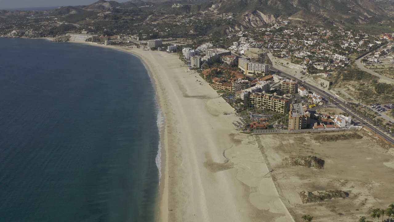 Aerial shoreline view with dark ocean, pale beach, resorts, road, and hills.