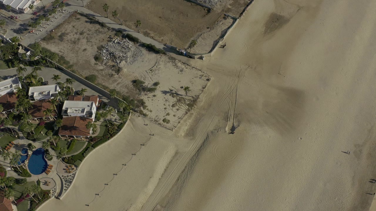 Aerial view over Baja California Sur, Mexico, showing pale beach sand, walled beachfront homes, palms, and a blue pool with no visible people.