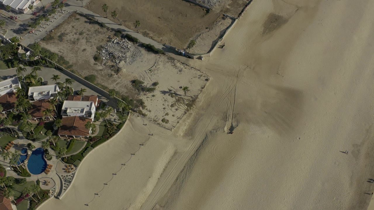 Aerial frame from Baja California Sur, Mexico, with beachfront houses and palms at left, wide pale sand at right, and no visible people.