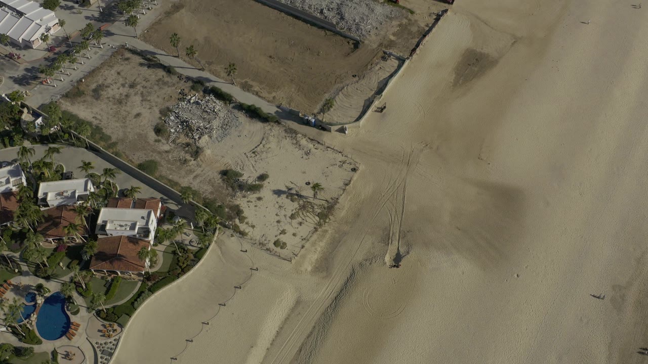 Aerial view in Baja California Sur, Mexico, showing walled lots, low homes, a pool, palms, and broad beach sand with no visible people.
